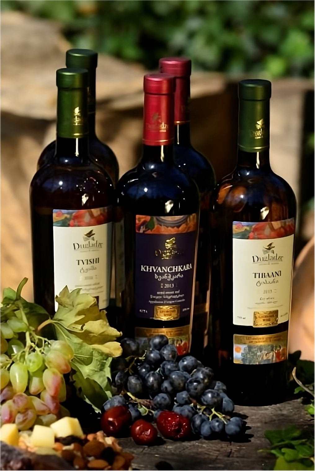 A selection of three Dugladze wine bottles labeled as "Tsinandali," "Kindzmarauli," and "Tsolikouri," surrounded by an assortment of fresh grapes and other fruits on a rustic table setting