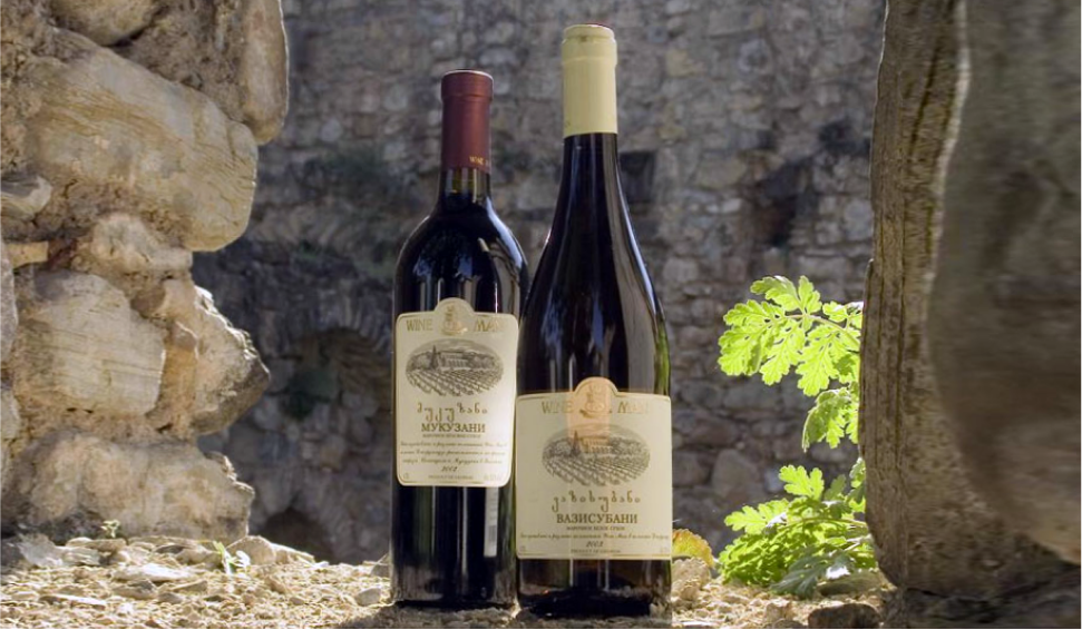 Two Wine Man bottles standing on a stone surface with an ancient stone wall and green foliage in the background