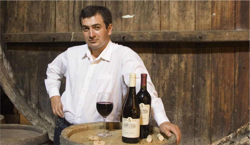 Wine Man Winery, Winemaker standing beside a wooden barrel with bottles of wine and a glass of red wine, in a wine cellar